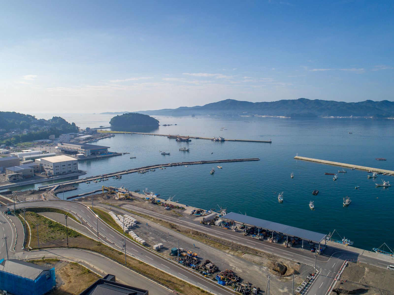 港のある海沿いの地域を上空から撮影したもので、水面には複数の船が浮かび、岸沿いには桟橋や建物が並び、背景には山々と島が広がり、空には雲が少し浮かび、手前には曲がりくねった道路や倉庫・工業施設のようなインフラが見える写真