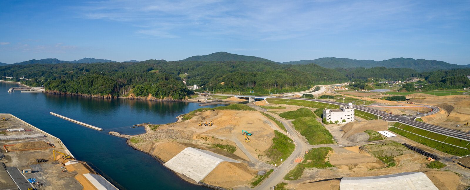 海沿いの地域で建設作業が進められている様子を上空から撮影したもので、左側に水面が広がり、周囲の陸地には舗装された道路や工事中の道路があり、中央右には白い建物を含む複数の構造物が見え、背景には緑の丘と森林が澄んだ青空の下に広がるパノラマ写真