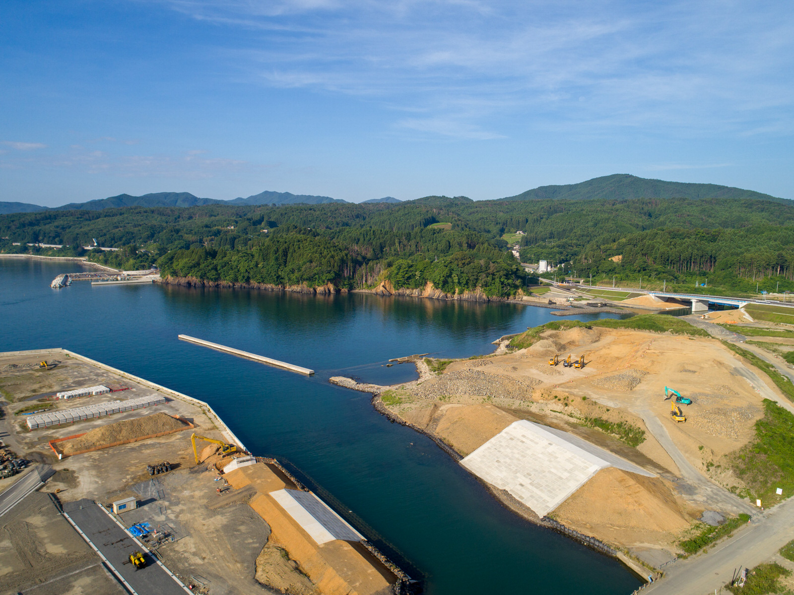 海沿いの建設現場を上空から撮影したもので、複数の掘削機などの建設機材がある土がむき出しのエリアが広がり、水辺にはコンクリート構造物や防護壁が見られ、背景には緑の丘と森林が広がり、空は雲が少なく晴れている写真