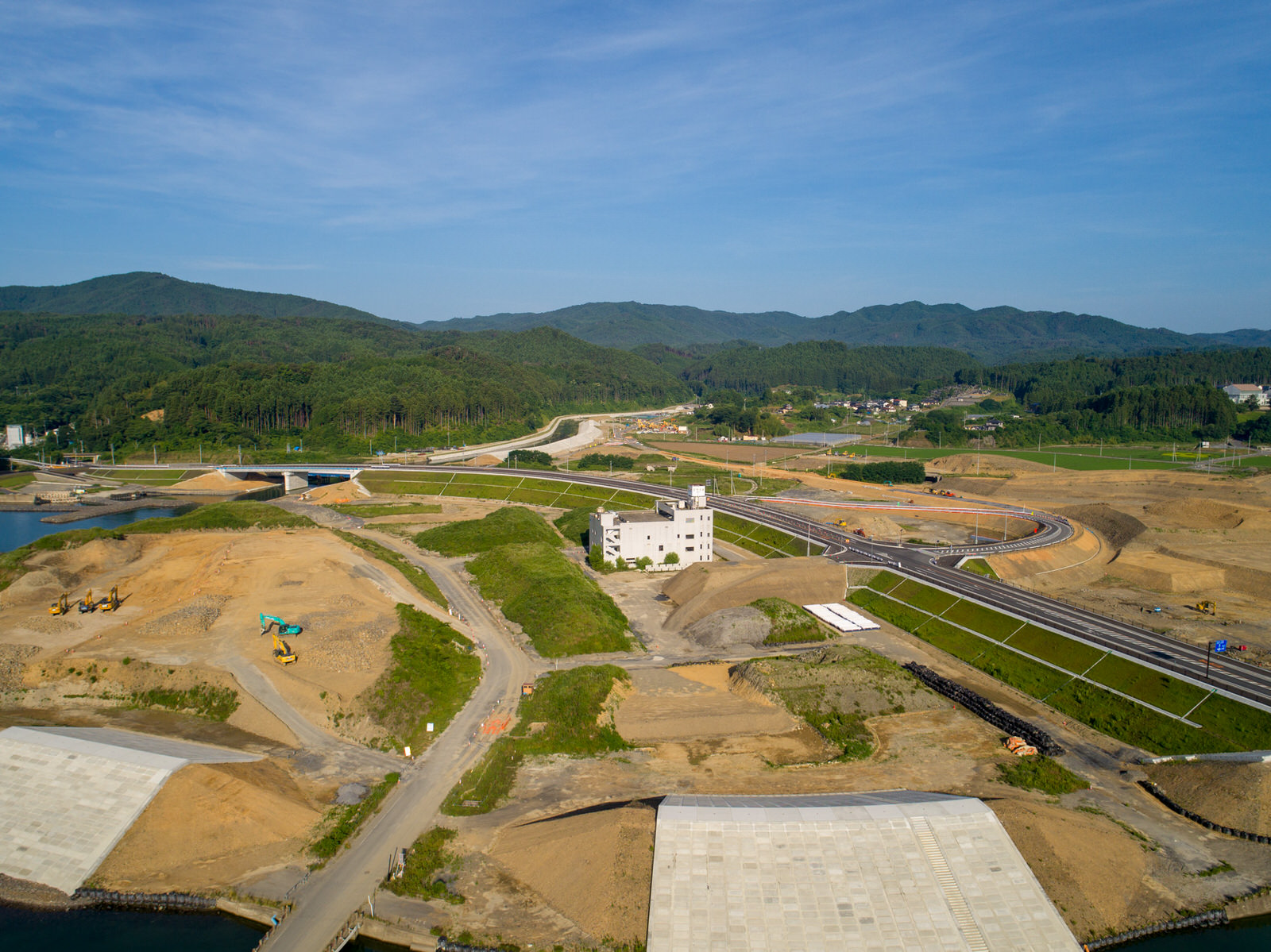 建設機材や道路、建設途中の建物がある工事現場を上空から写した風景で、周囲には緑の丘と山々が広がる写真