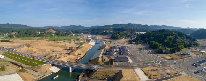 川が中央を流れる田舎の風景で、複数の橋が川を横断し、両岸で建設作業が行われており、背景には緑の山々が広がり、建物や道路、駐車場も見えるパノラマ写真