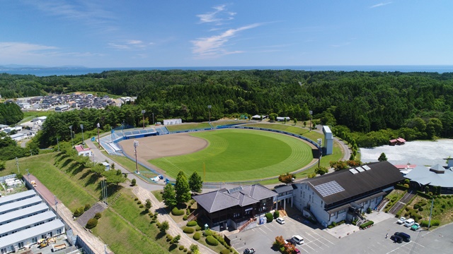 緑豊かな森に囲まれた丘の上に位置する円形の野球場があり、晴れた青空の下で周囲に駐車場や建物が整備されている風景を上空から撮影した写真