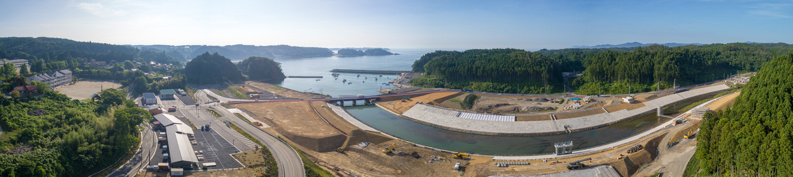 中央に広がる水面の周囲で開発が進む海沿いの地域を広く見渡した風景で、建設車両や機材、新しく整備された道路や構造物が見られ、背景には緑豊かな森林と丘、そして水平線まで続く海が広がるパノラマ写真