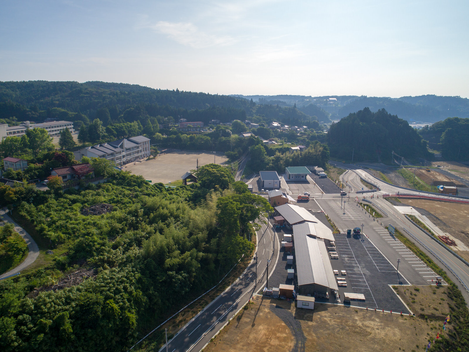 建物や道路、緑が混在する地域を上空から撮影したもので、倉庫や商業施設と思われる構造物と駐車場、広大な敷地に建つ校舎が見える学校があり、周囲には木々や草地が広がり、背景には丘が見える写真