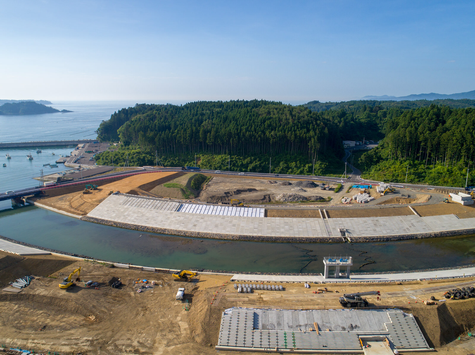 川や運河のそばにある建設現場を上空から撮影したもので、複数の掘削機やトラックなどの建設車両が見られ、水辺には大きなコンクリート構造物や擁壁が並び、背景には密集した森林とその奥に広がる丘や山々が澄んだ青空の下に広がる写真