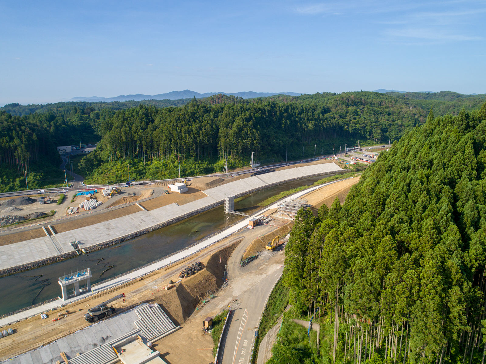 森林に囲まれた地域の建設現場を上空から撮影したもので、中央には川が流れ、両岸にはコンクリートの護岸が整備され、周囲には建設機材や資材が並び、背景には密集した森林と丘が澄んだ青空の下に広がる写真