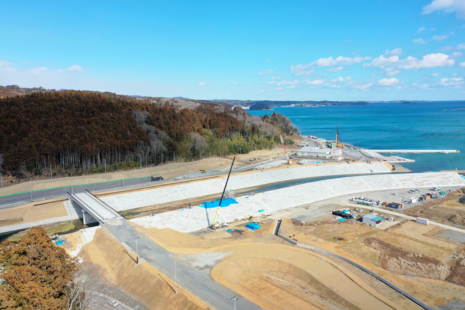 海沿いの工事現場で大型クレーンが稼働しており、周囲には山と海が広がっている様子が一望できる風景の写真