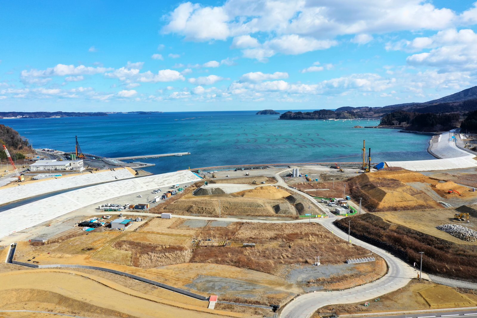海沿いの工事現場で、複数のクレーンや建設車両が稼働し、大きな土の山や建設中の構造物が点在し、背景には青く澄んだ海と雲のある空が広がっている様子を捉えた開発中の風景の写真