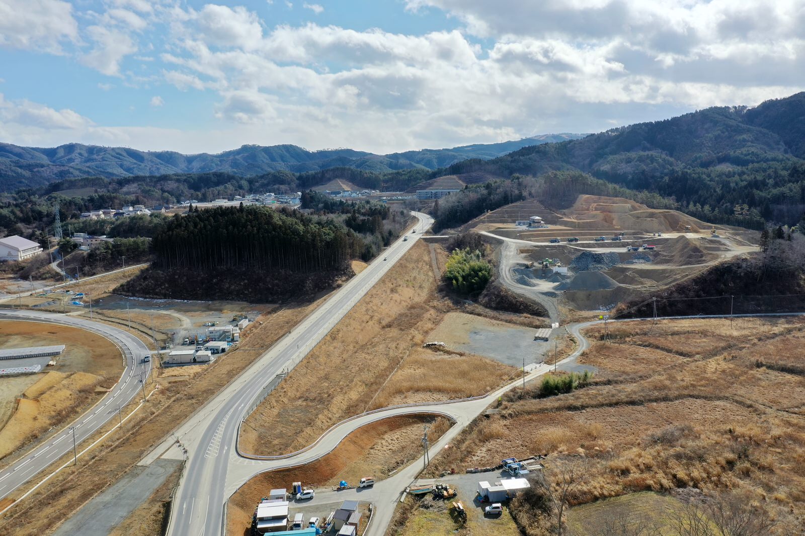 丘陵地帯を切り開いて新しく整備された道路が伸びており、その沿道には複数の工事車両や機材が見られ、周囲には森林や遠くに建物が点在し、空には雲と青空が広がっている風景の写真
