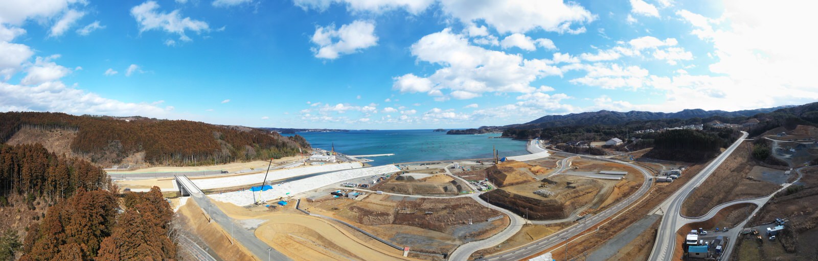 海に面した広大な工事現場が広がり、道路や通路、建物などが整備中で、背景には海とそれを囲む丘や森林が見え、空には少し雲が浮かんでいる様子を一望できる風景の写真