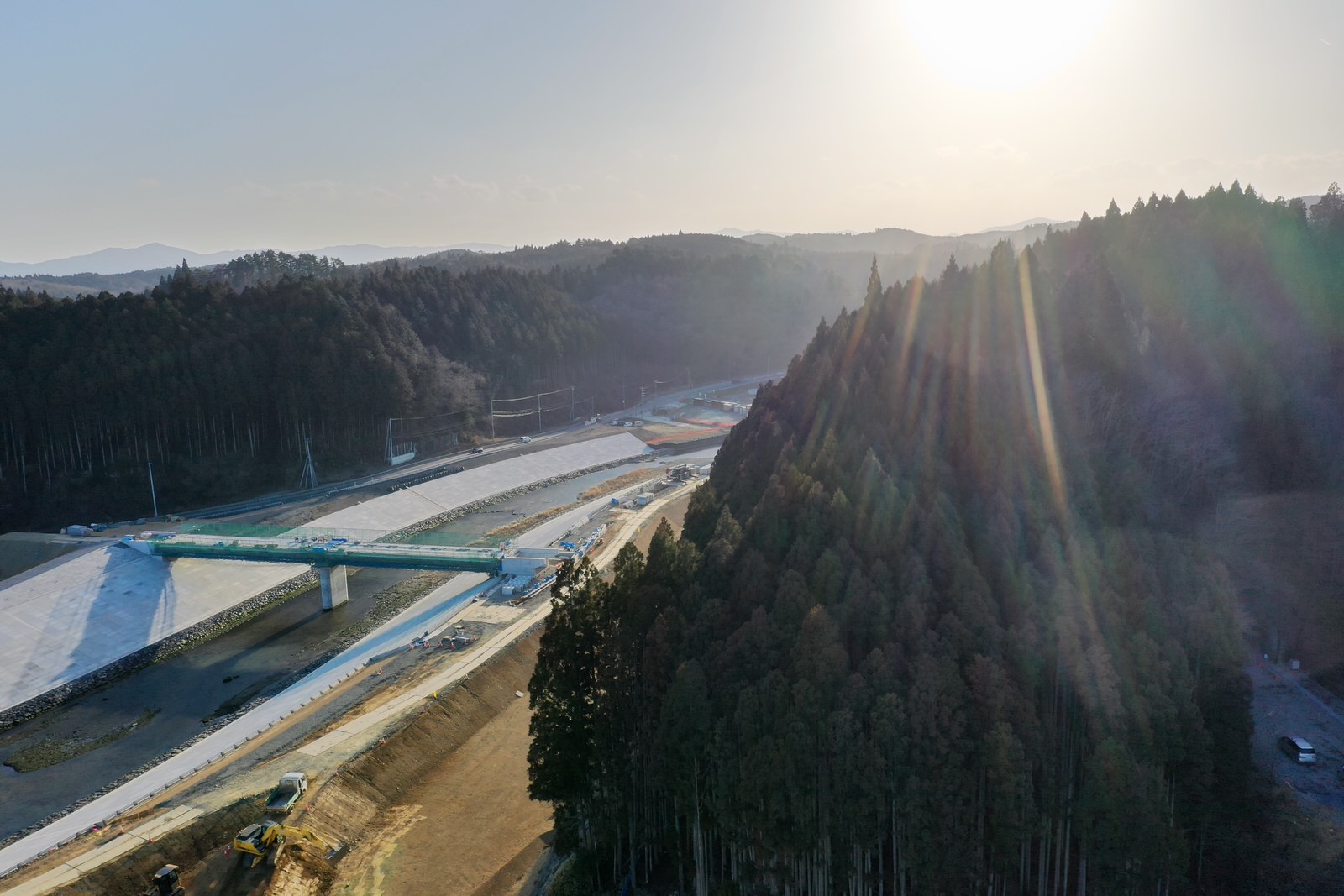 川に架かる橋とその周辺に広がる森林に囲まれた山間の風景で、太陽の光が明るく差し込み、川岸には工事車両や機材が見られる自然と開発が共存する地域の写真