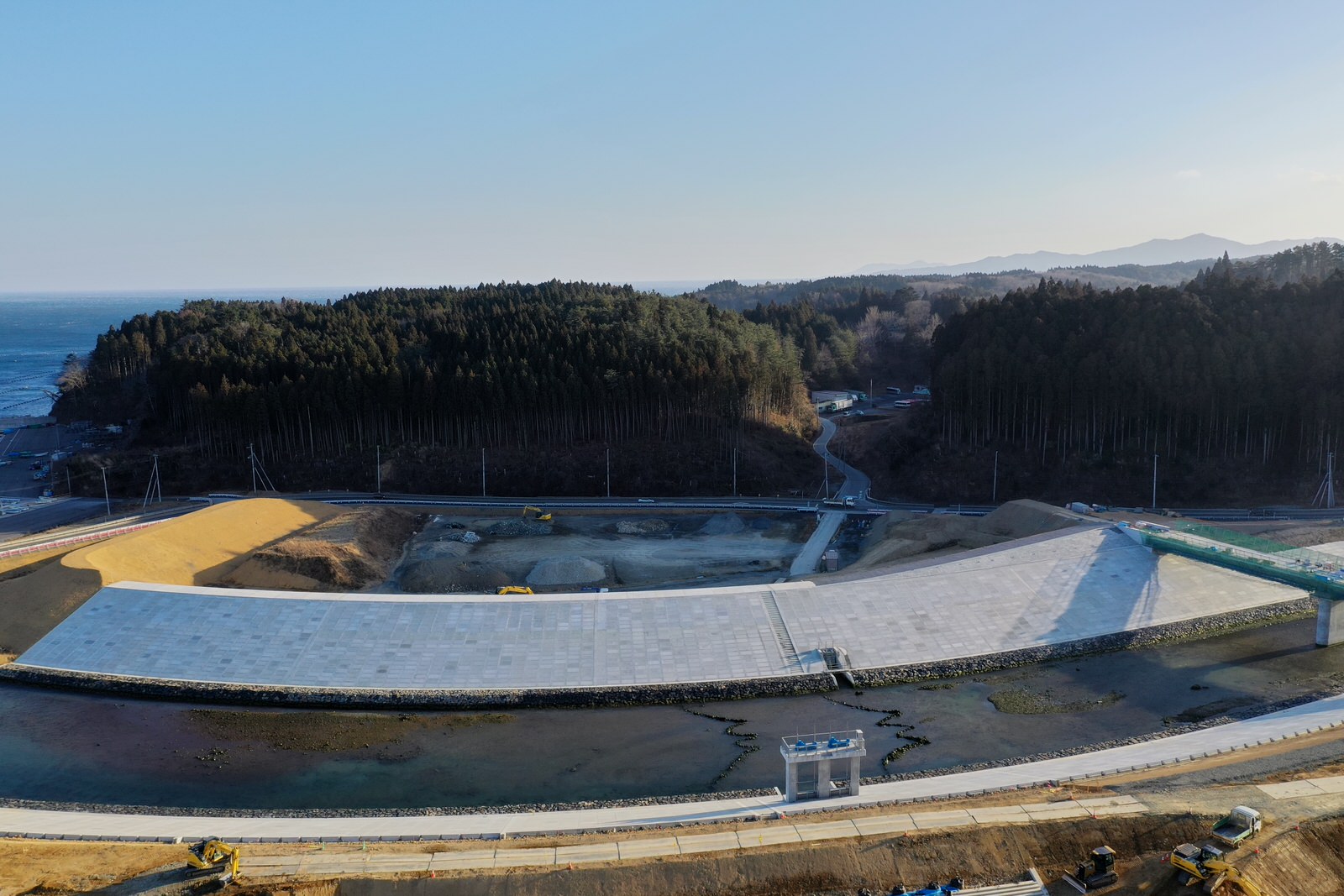 海岸沿いに大きく築かれた防波堤が手前にあり、その背後には緑豊かな森林が広がっている自然と人工構造物が共存する風景の写真