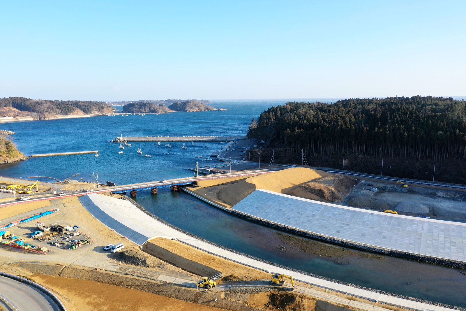 海に面した工事現場で、新しく整備された堤防や土木構造物が広がり、複数の重機やトラックが稼働しており、背景には青い海と小さな船、両岸には森林が広がる晴天の風景の写真