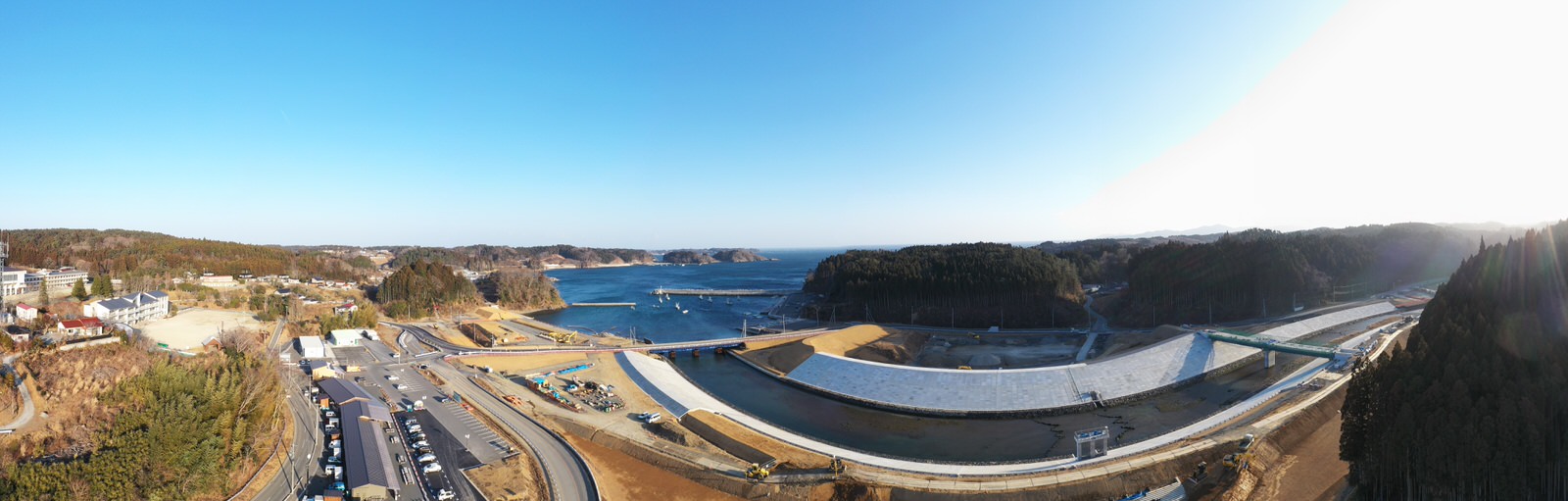 海に面した広大な工事現場で、湾曲したコンクリート構造物が風景を横切り、周囲には道路や駐車された車両、建物が点在し、背景には木々に覆われた丘と遠くに海が広がる晴天の風景の写真