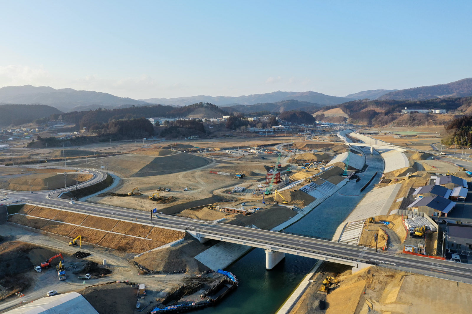 川または運河に架かる橋を中心に、複数の工事車両や資材が並ぶ大規模な建設現場が広がり、周囲には整備中の道路や通路、右側にはいくつかの建物が見え、背景には山々が連なる風景の写真