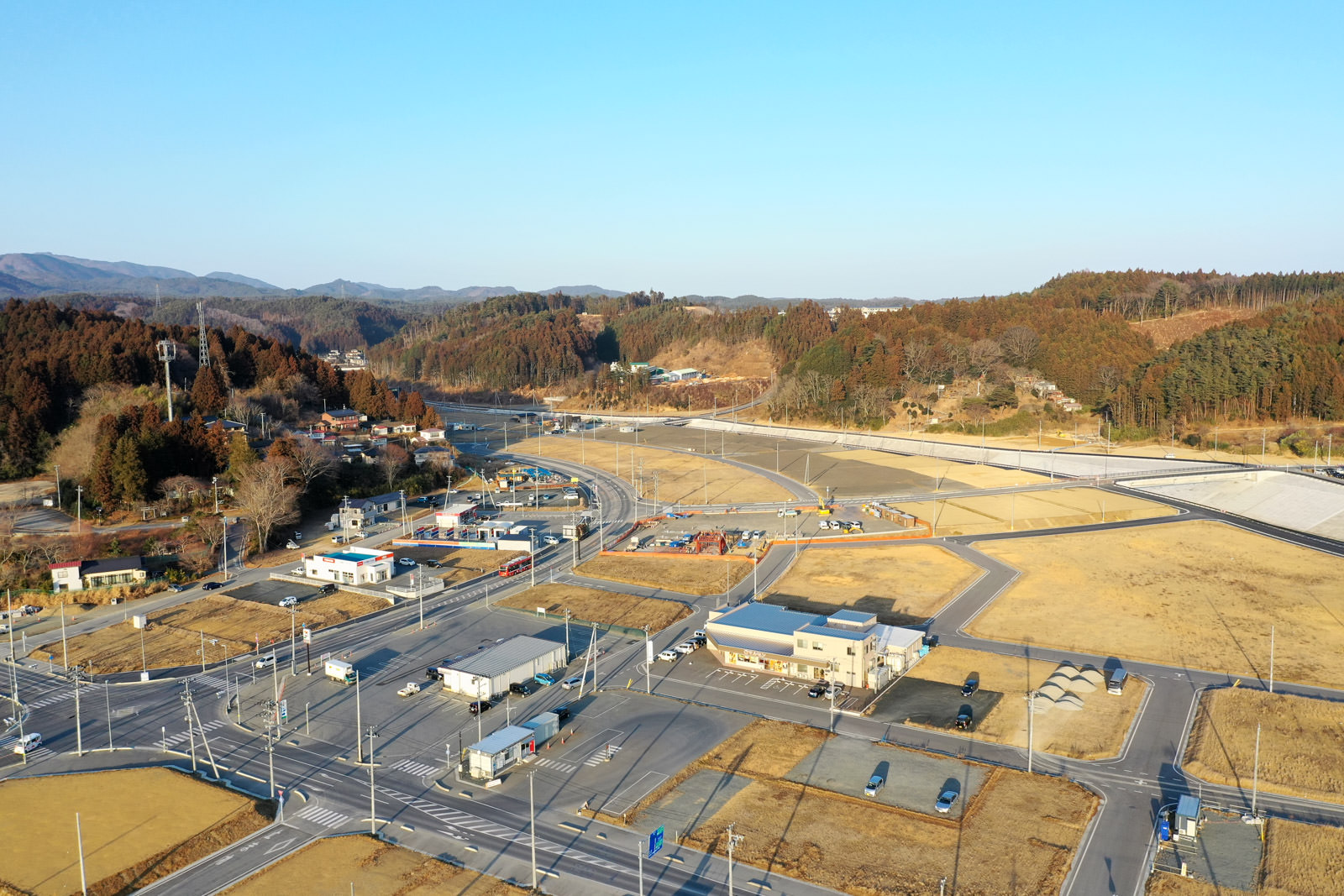 広々とした道路と交差点が田舎の風景の中にあり、周囲には山や木々が広がり、いくつかの建物や車も見える静かな地域の写真