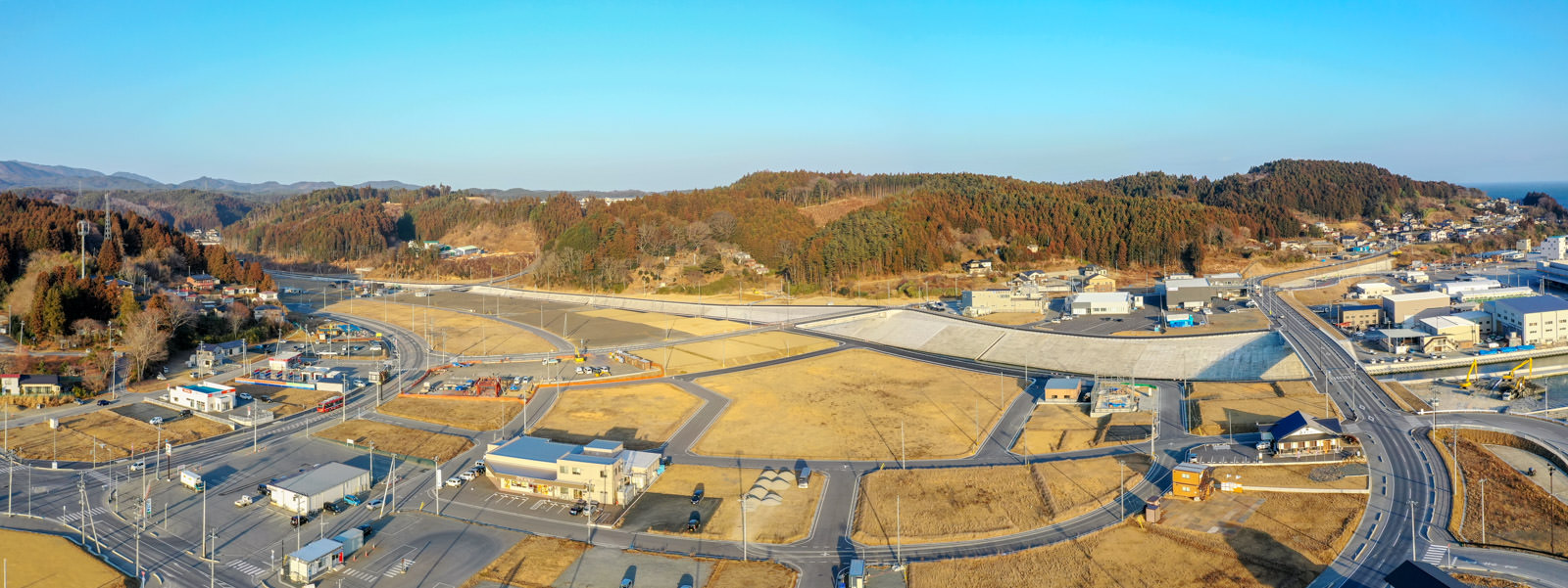 住宅と未開発の土地が混在する農村地域を空から見下ろした写真で、複数の家屋や道路、広い空き地が広がり、背景には木々に覆われた丘と澄んだ青空が見える田園風景の写真