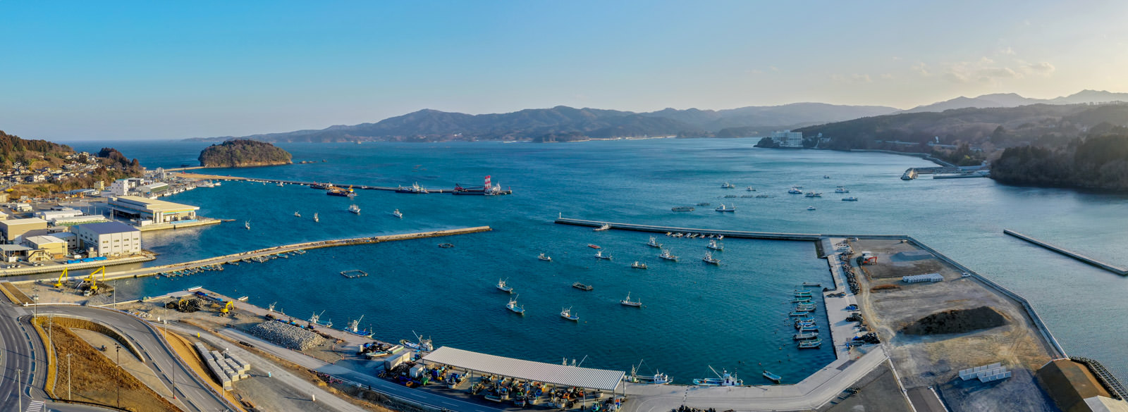 三方を陸地に囲まれた海辺の港に複数の船が停泊しており、岸沿いには建物や施設が並び、背景には丘や山が広がり、空には少し雲が浮かんでいる様子を一望できる港の写真