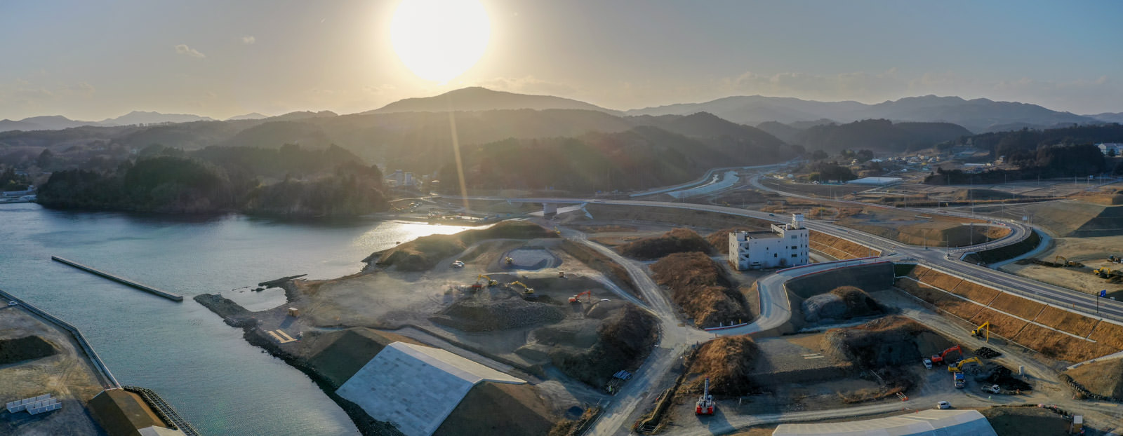 山の上に昇る太陽が空を明るく照らし、手前には静かな湖が広がり、その周囲には道路や建物が点在している自然と人の営みが調和した風景の写真