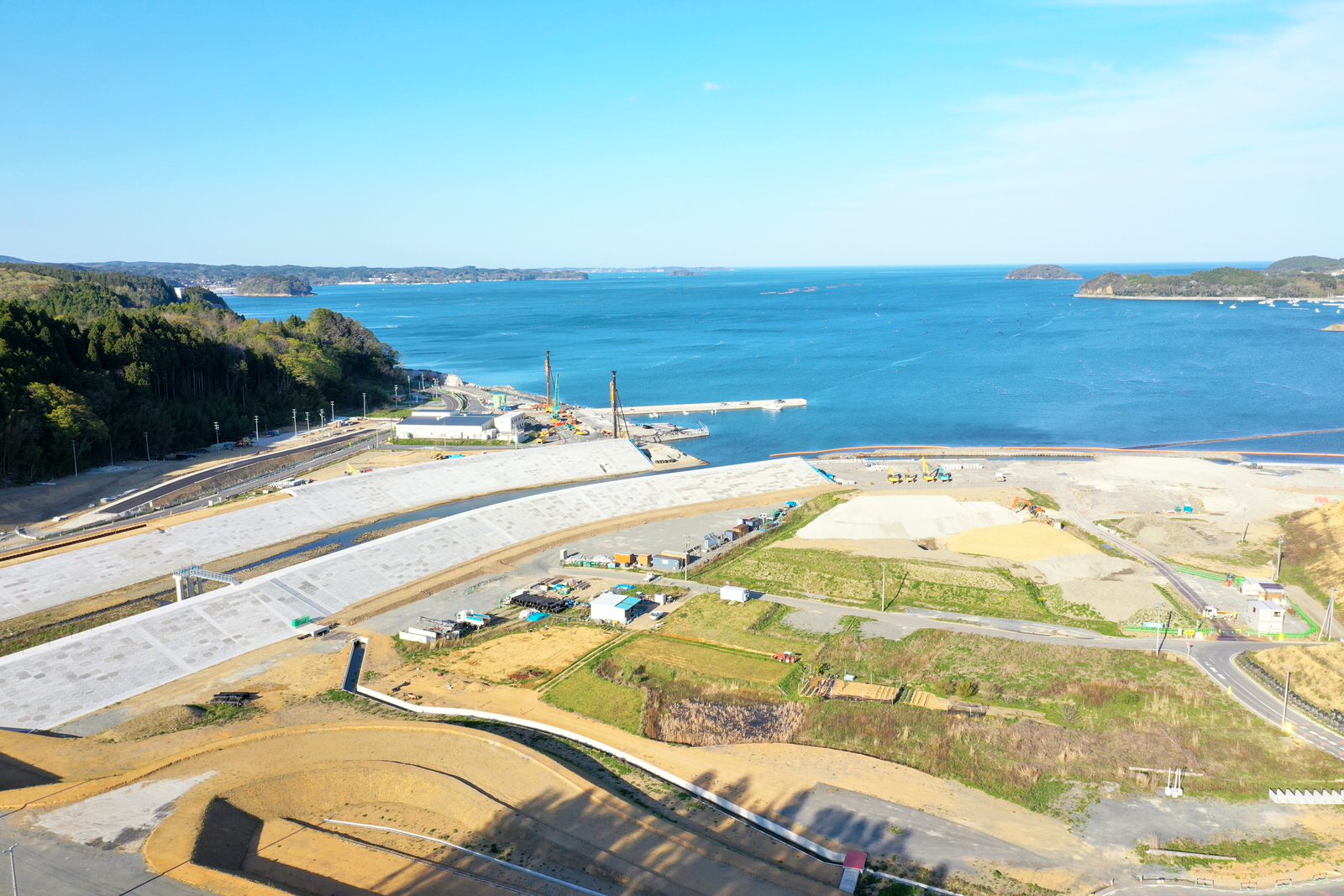 海岸沿いの広い敷地にコンクリートの基礎が整備され、海に向かってクレーンが立ち並ぶ工事の様子を空から撮影した海辺の工事現場の写真