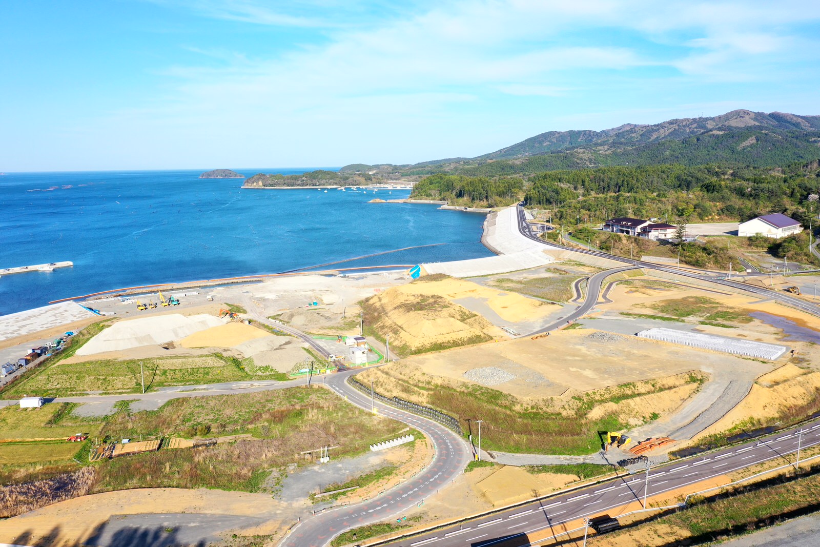 海岸沿いの広い土地に道路が整備され、建設工事が進行する様子を空から撮影しており、背景には青い海と山々が広がる海辺の開発地域の写真