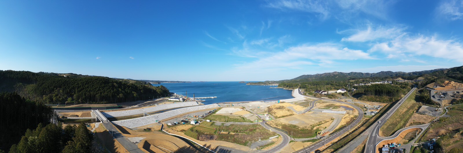 青い空の下、海に面した広い土地に道路や建設中の施設が点在し、自然と開発の様子が共存する風景を空から撮影した海沿いの開発地域の写真