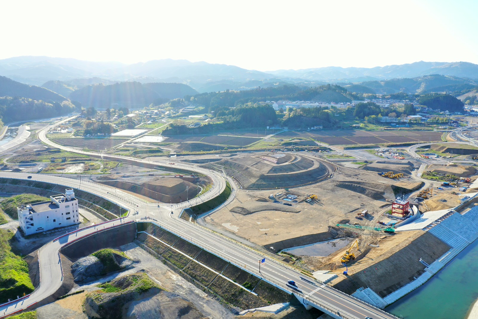 山間部の広大な土地に複数の道路が交差し、建設中のインフラや建物が点在する様子を空から撮影した山間の開発地域の写真