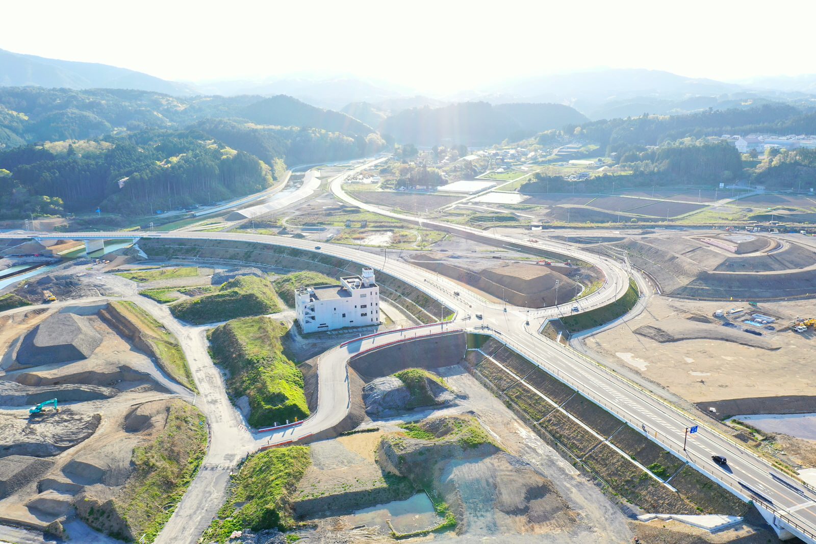 山間部にある道路工事現場を空から撮影しており、周囲には森林が広がり、重機や資材が点在する様子が確認できる道路工事現場の写真