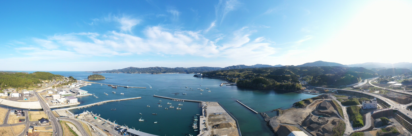 青い空の下、海に囲まれた港町に船や建物、道路が点在し、自然と人の営みが調和する様子を空から撮影した港町の写真