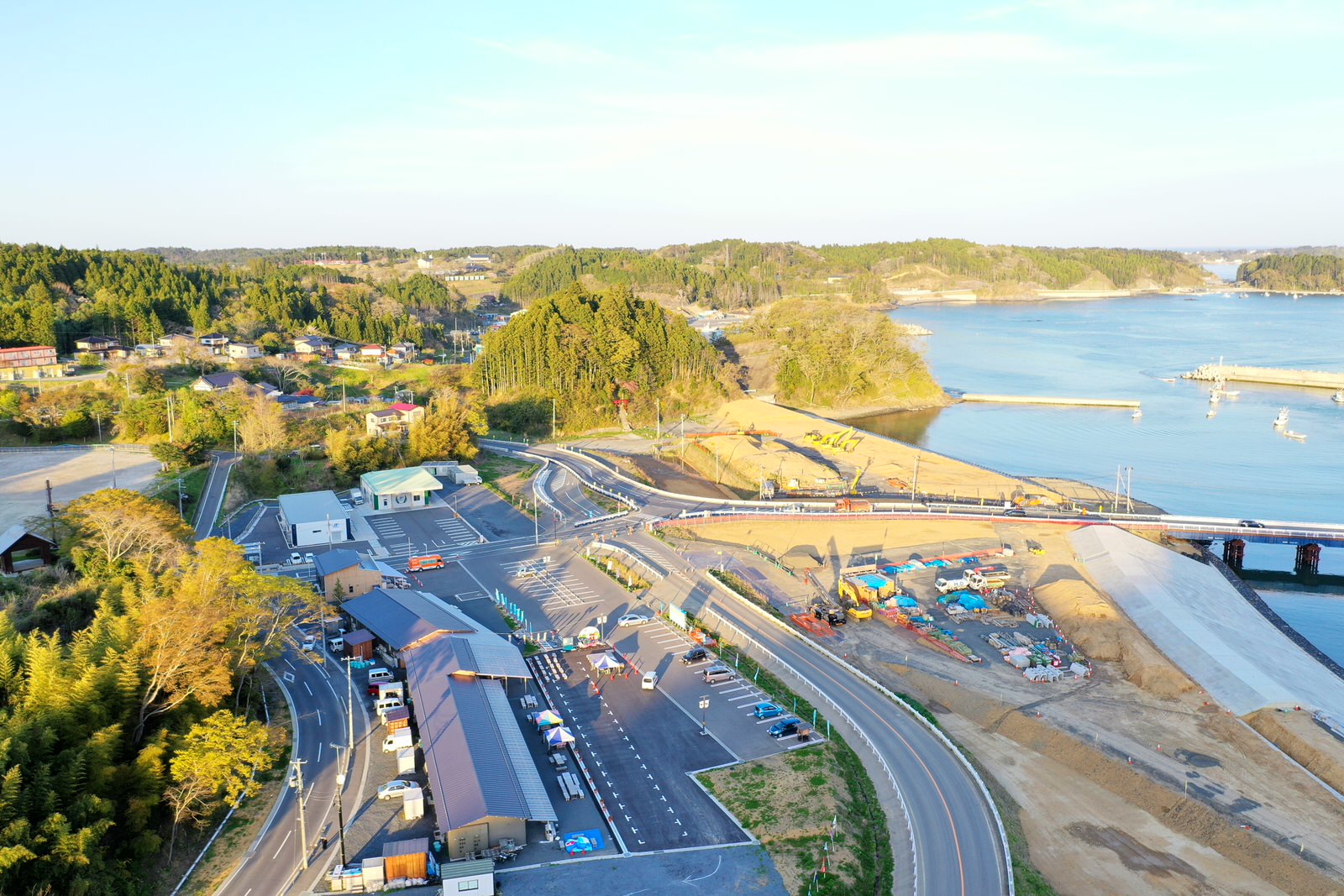 手前に広い駐車場と複数の建物が並び、奥には川が流れ、その上に橋が架かっている海沿いの町を上空から撮影した町の写真