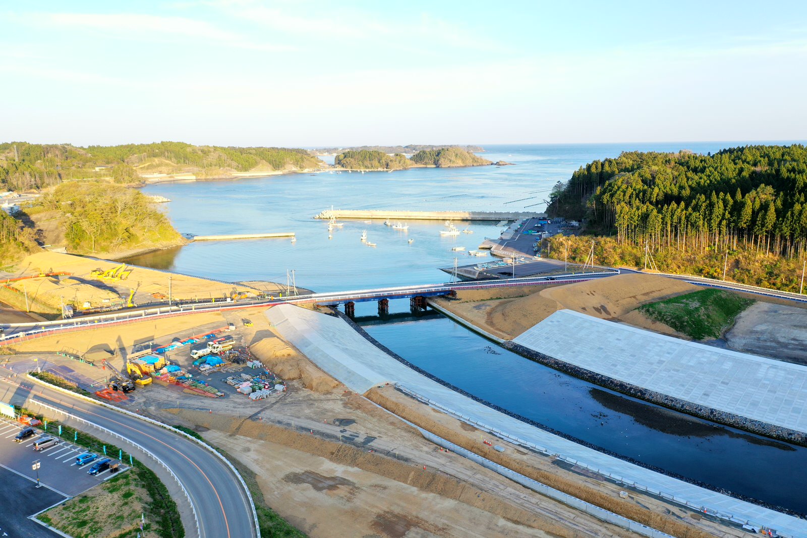 森林に覆われた丘に囲まれた海沿いの地域を上空から撮影した写真で、川が海へと流れ込み、その川には橋が架かっており、両岸では工事が行われていて、小さな港には船が停泊している晴天の海辺の写真