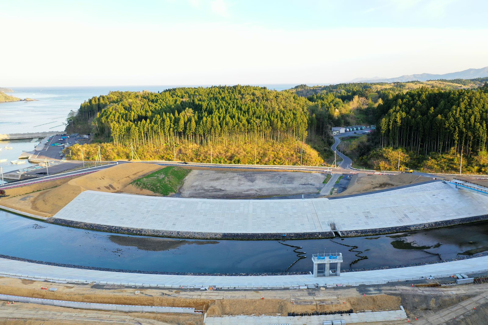 手前に広がる水面の奥に防潮堤の構造物があり、その背後には密集した森林と一部開けた土地が広がり、周囲には構造物から森へと続く道路や小道が見え、左奥には海、右奥には丘陵地が広がる風景の写真