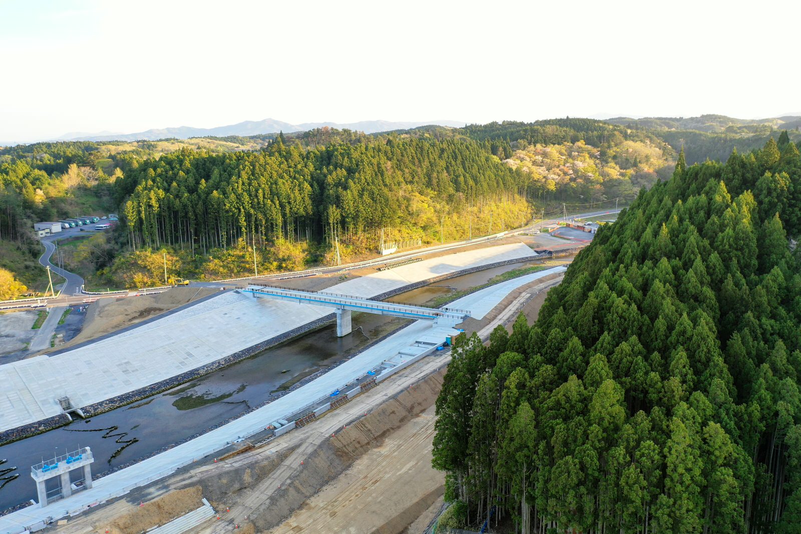 森林に囲まれた地域で川にかかる橋の建設現場を写した写真で、コンクリートの構造物や支柱が見える新しく建設中の橋が中心にあり、周囲には密集した森と工事のために開かれた土地、そして丘陵地が広がる橋の工事現場の写真
