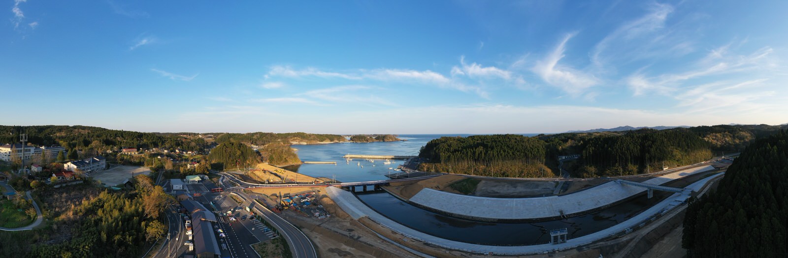 青空の下、海に面した小さな町とその周辺の風景が広がり、手前には道路や建設中のインフラが点在する様子を空から撮影した海辺の町の写真