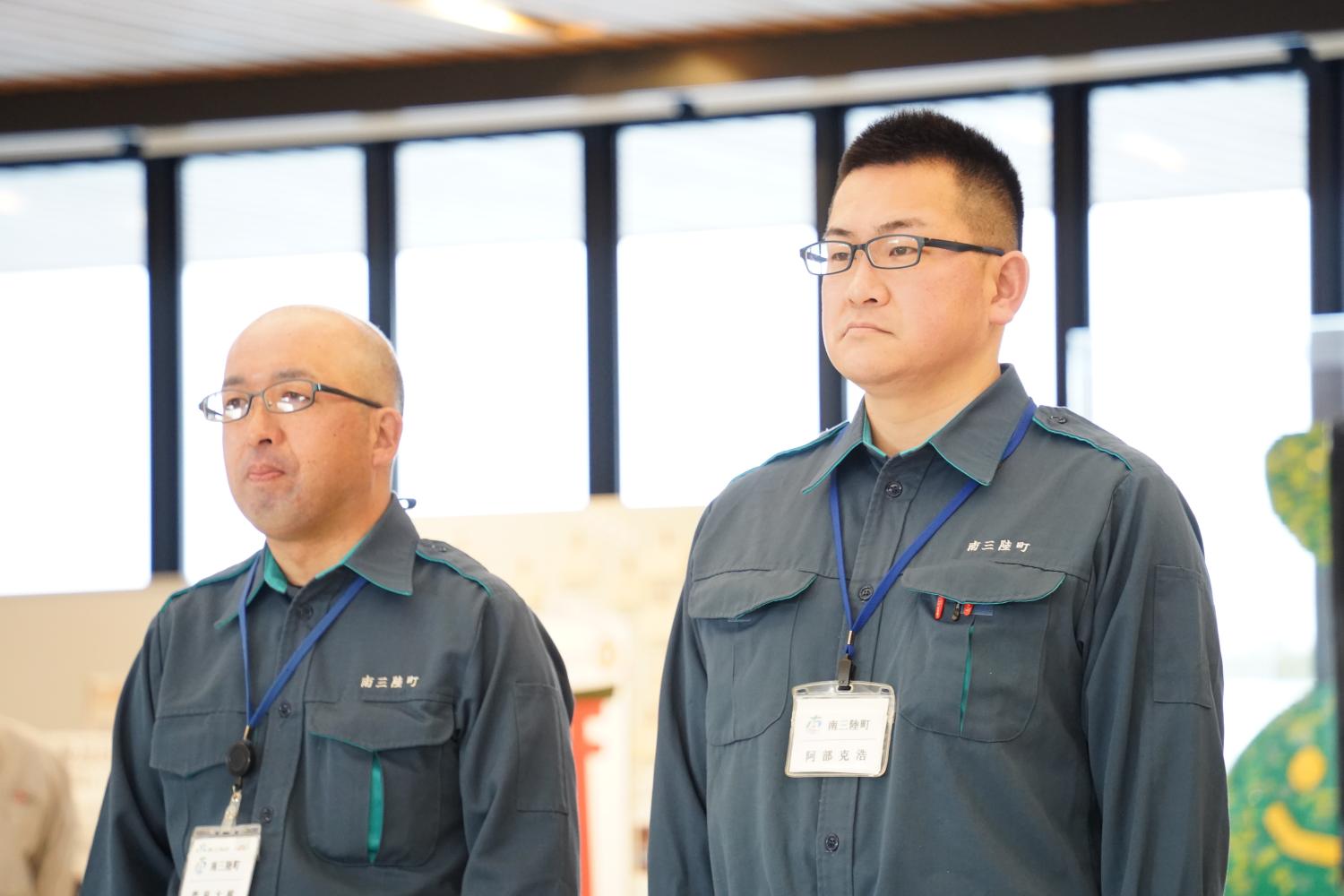 作業服を着た2人の男性が名札を首から下げており、真剣な表情で前を見ている写真