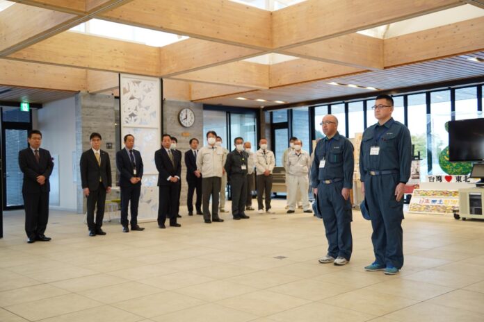 関係者に見守られ、中央には作業服を着た2人が前に立ち出発式が行われている様子の写真