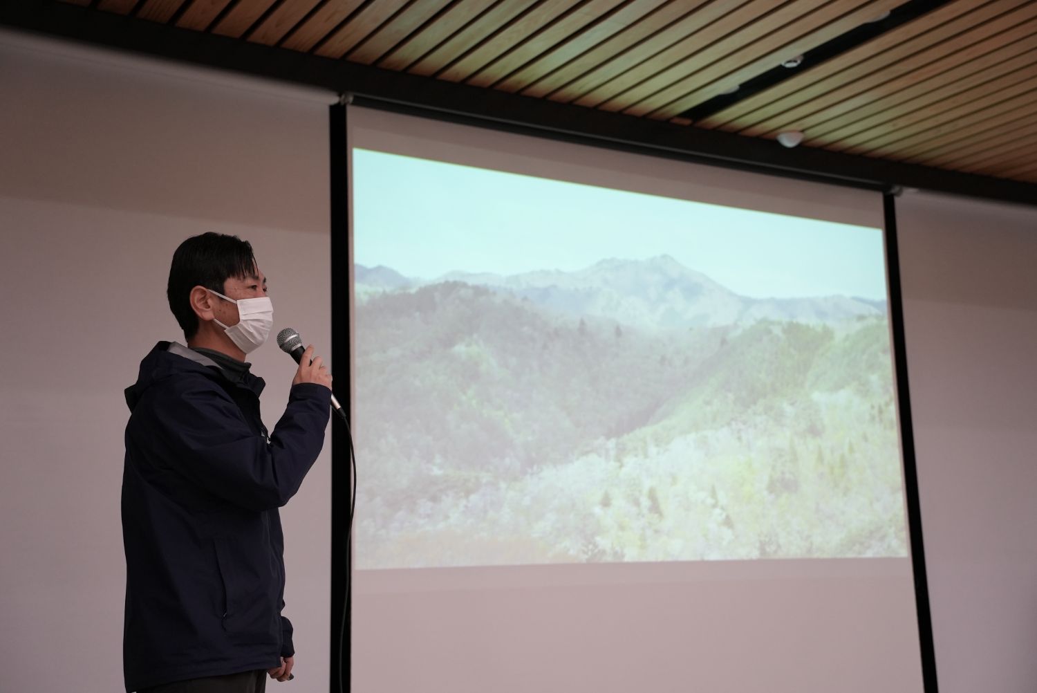 白いマスクとネイビーのジャケットを着た鈴木卓也さんが山の風景が映されたスクリーンの前でマイクを持ち、環境や自然保護について語っている様子の写真