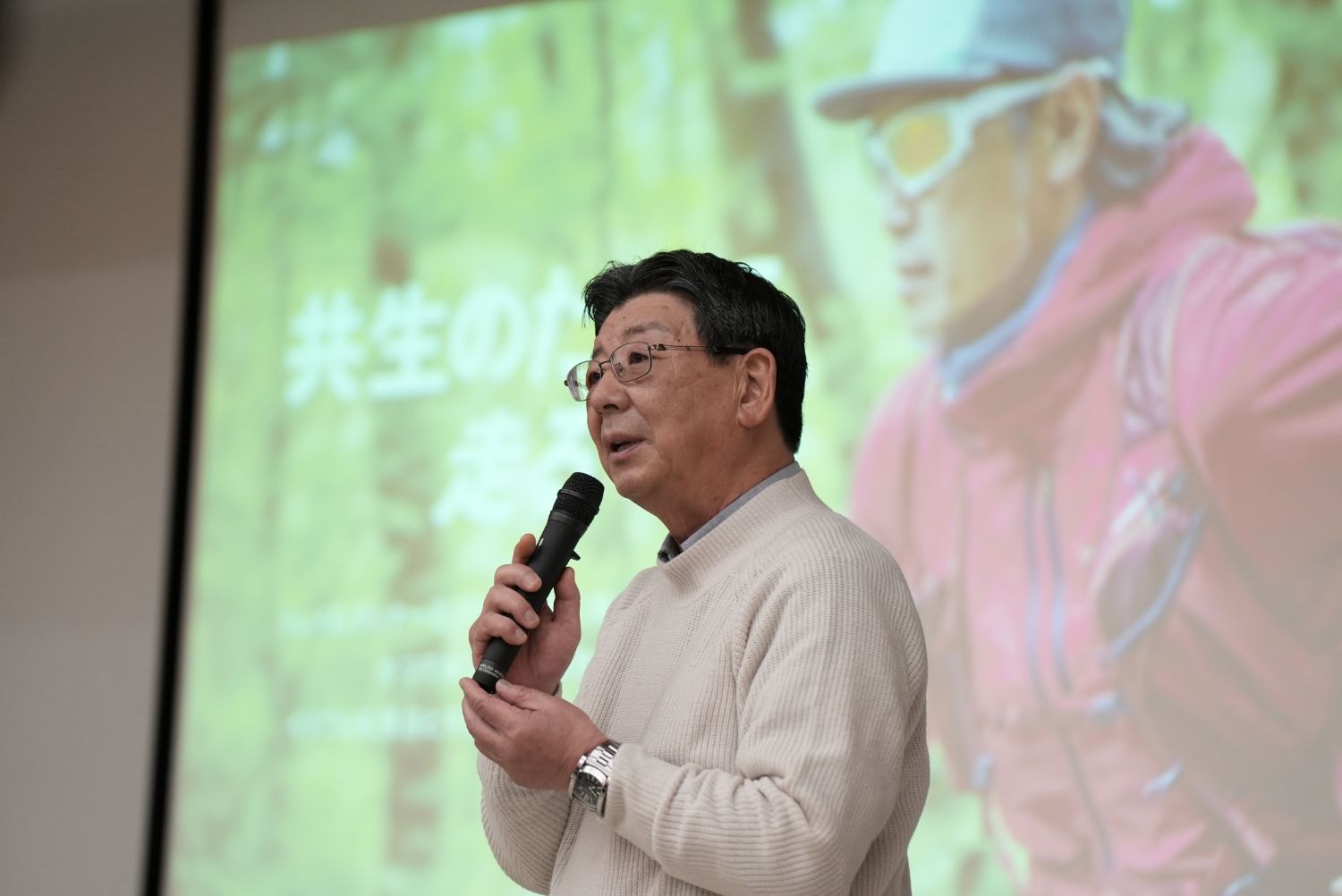 薄いベージュのセーターを着た佐藤仁町長が、森林を背景に「共生のために走る」と表示されたスクリーンの前でマイクを持って真剣に話している様子の写真