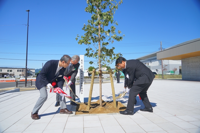 晴天の下、スーツ姿の男性3人が、白いタイル敷きの広場で若木を囲み、赤白のリボンが付いたスコップで丁寧に土をかけて植樹を行っている写真