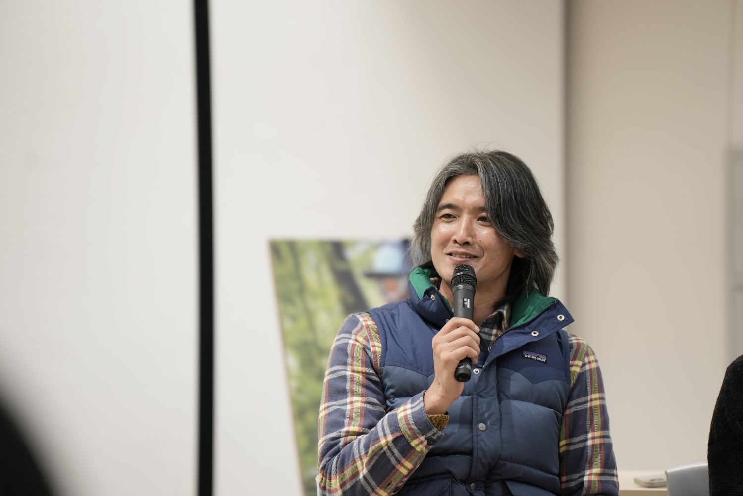 チェックのシャツに青いダウンベストを重ねた石川さんが、マイクを手に笑顔で話をしている様子の写真