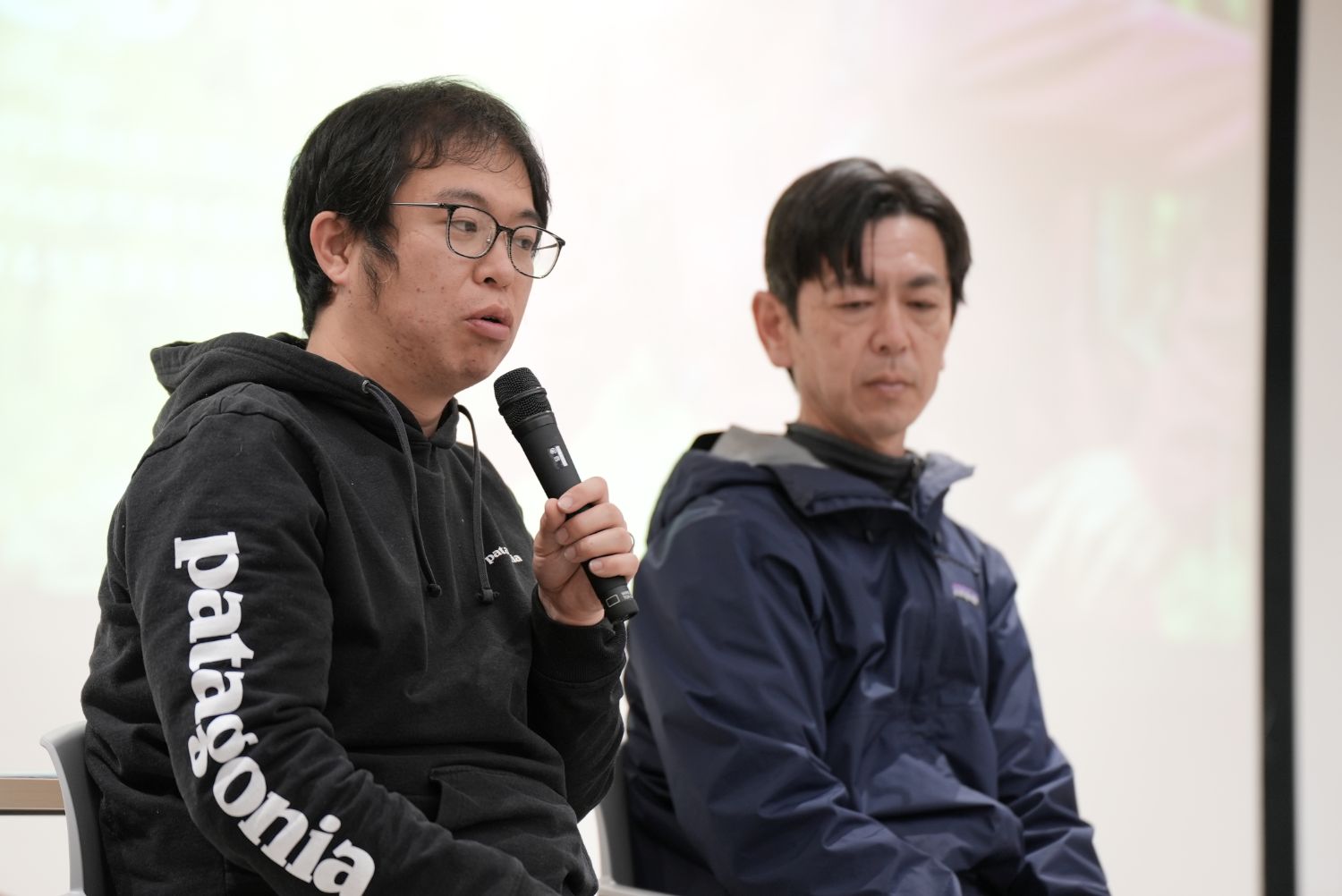 パタゴニアのロゴ入りパーカーを着た若い男性がマイクを持って発言し、その隣でネイビーのジャケット姿の男性が真剣な表情で耳を傾けており、環境共生についての意見交換が行われているトークイベント中の様子の写真