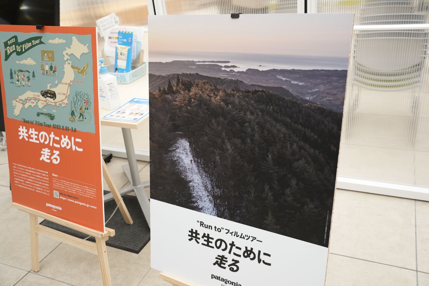 「共生のために走る」と書かれたフィルムツアーの2種類のポスターが並べて立てかけられており、左側は日本地図とイラスト、右側は森と海岸線の広がる風景写真が使われている展示の様子の写真
