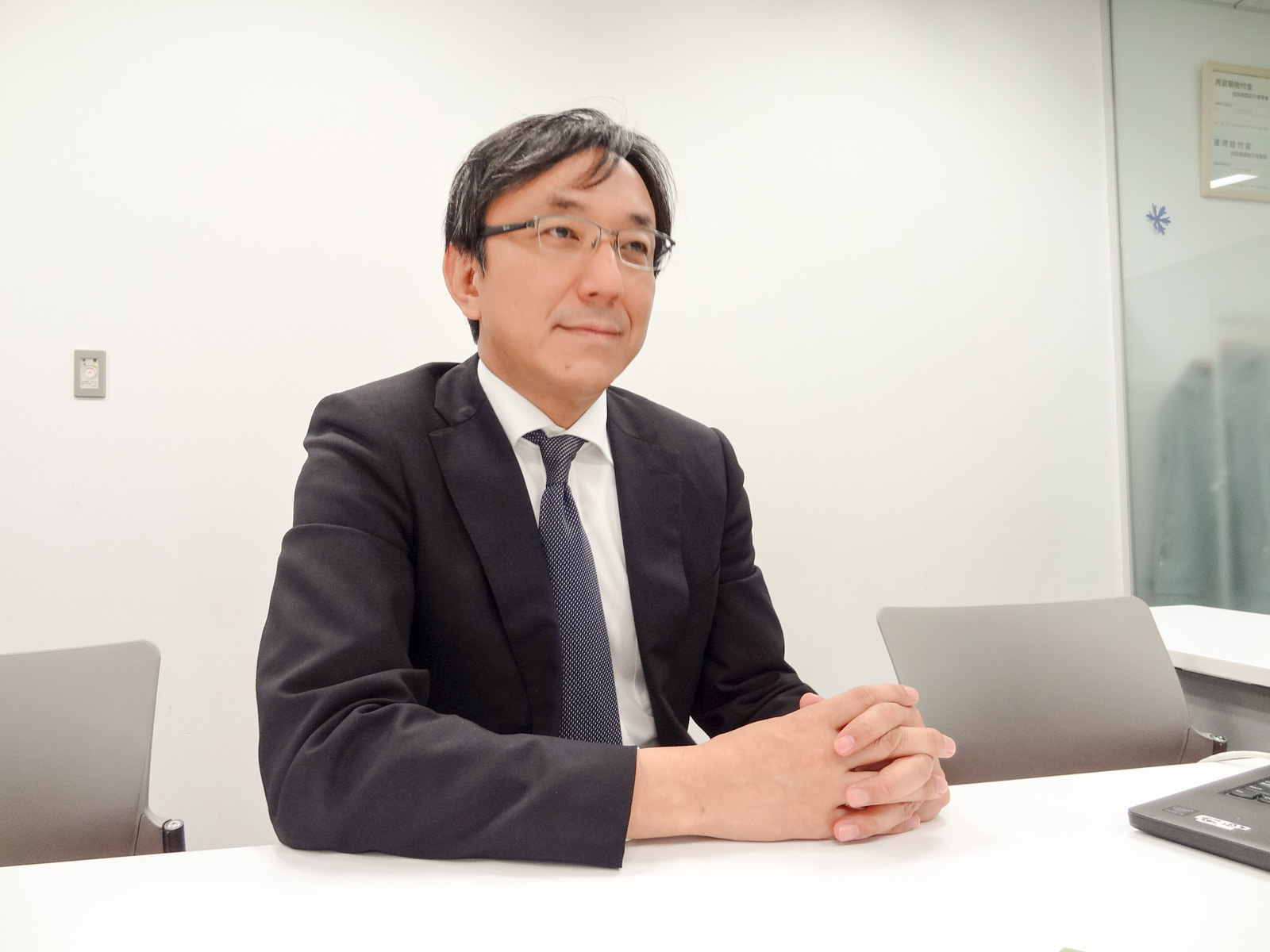 眼鏡をかけたスーツ姿の男性が白い会議机に両手を組んで置き、まっすぐ前を見ている様子の写真