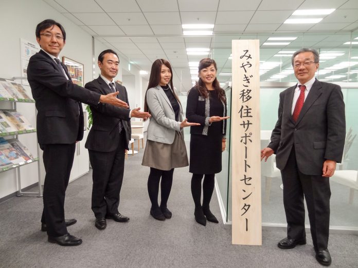 オフィスの中で「みやぎ移住サポートセンター」と書かれた縦長の看板の左右にスーツを着た男性3人と女性2人が立っている写真