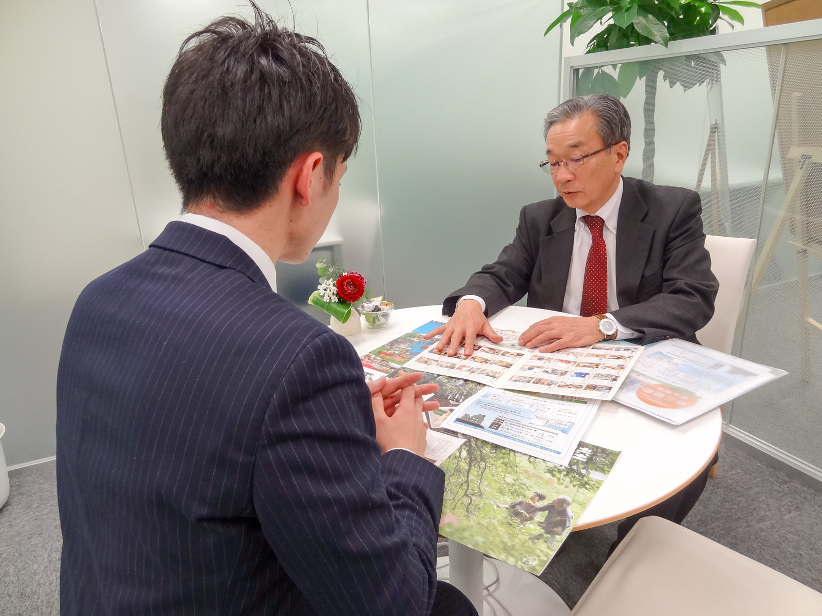 グレーのスーツを着た年配の男性が、紺色のスーツを着た若い男性と向かい合って座りながら、複数のパンフレットを机に広げ、説明をしている写真