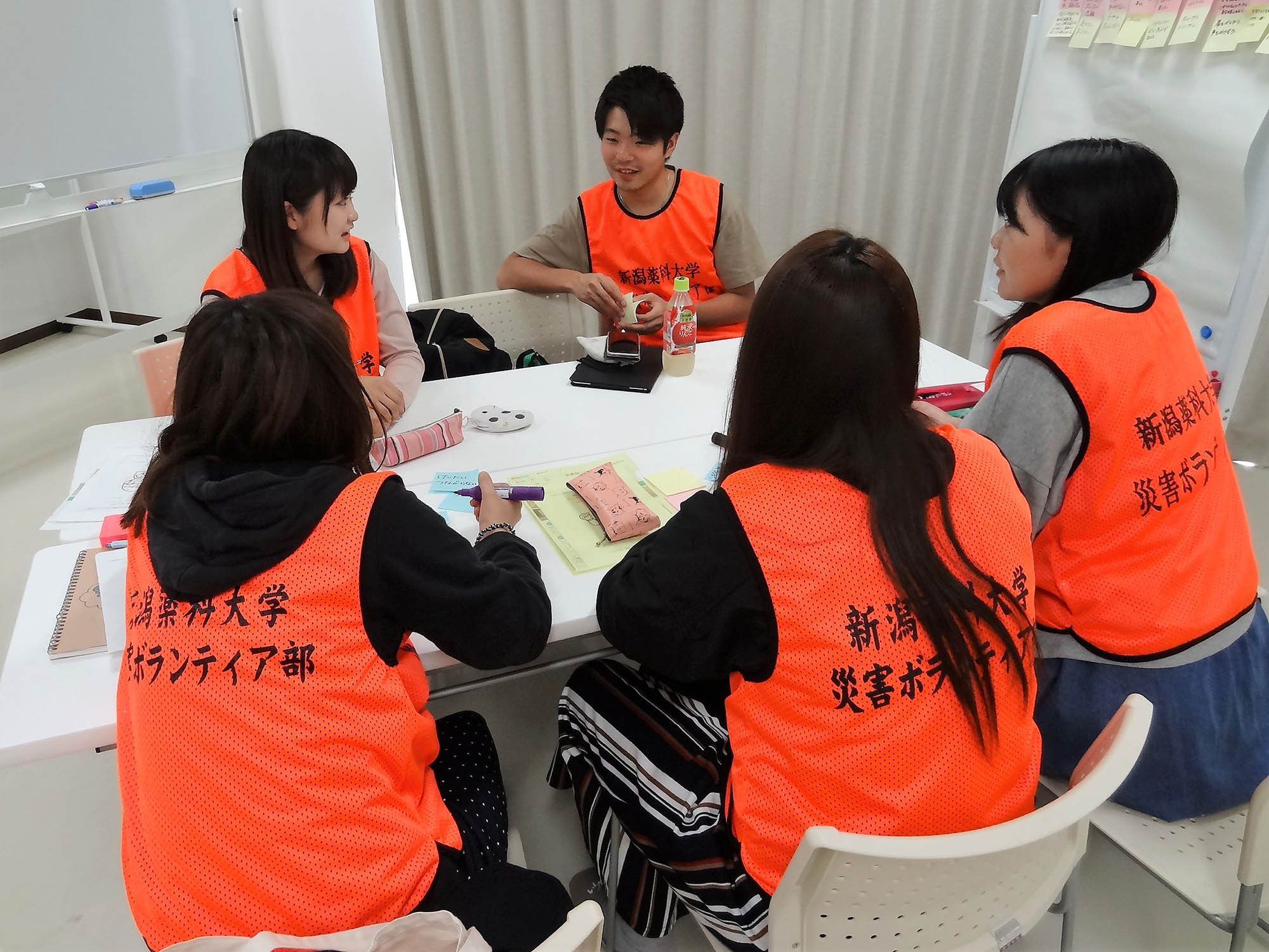 新潟薬科大学 災害ボランティア部』と書かれたオレンジ色のビブスを着た5人の大学生が、机を囲んで座り、災害に関する意見を付箋に書いたり話し合ったりしているミーティングの様子の写真
