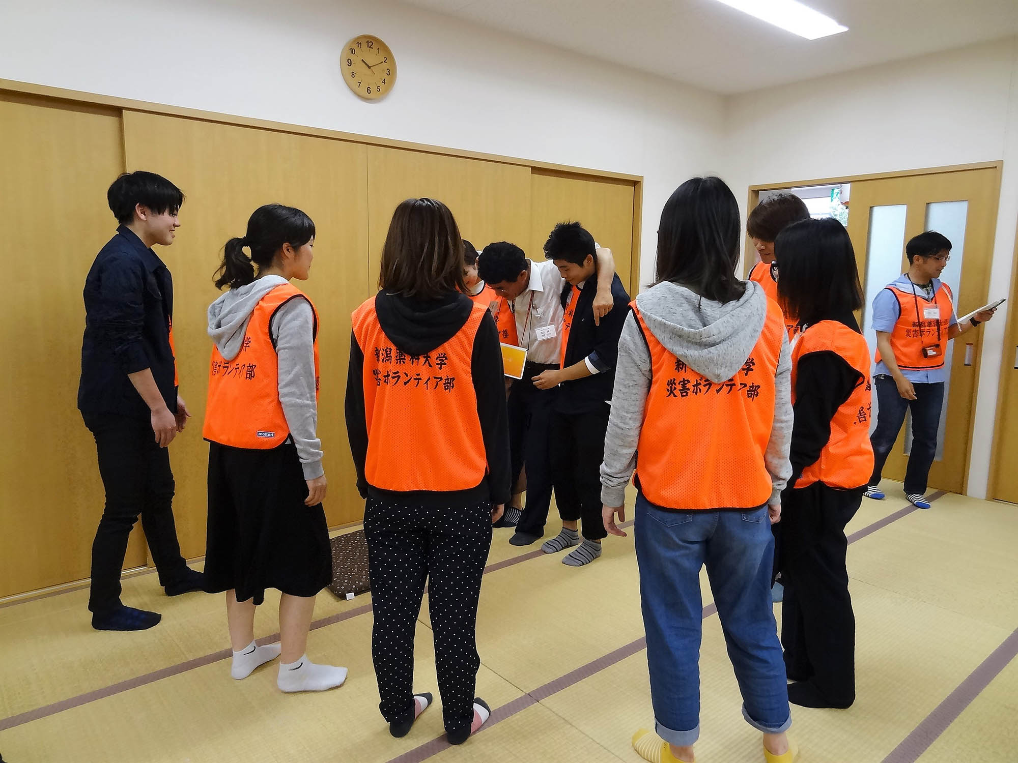 10人ほどの大学生がオレンジ色のベストを着て輪になって、けが人役を支えながら応急搬送の訓練をしているのを見学している様子の写真