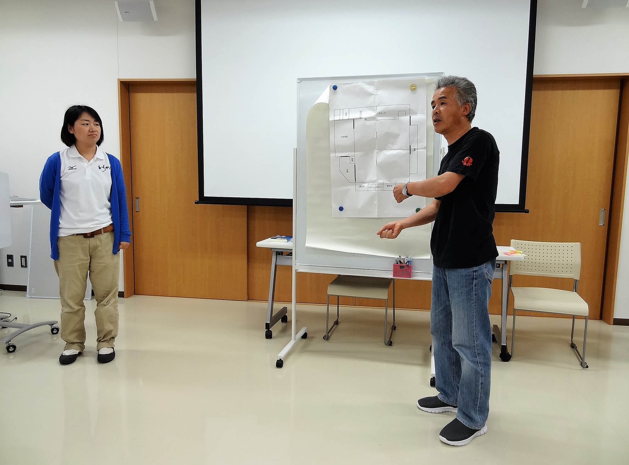 阿部忠義さんがホワイトボードに貼られた建物の間取り図を指差しながら話をしている写真