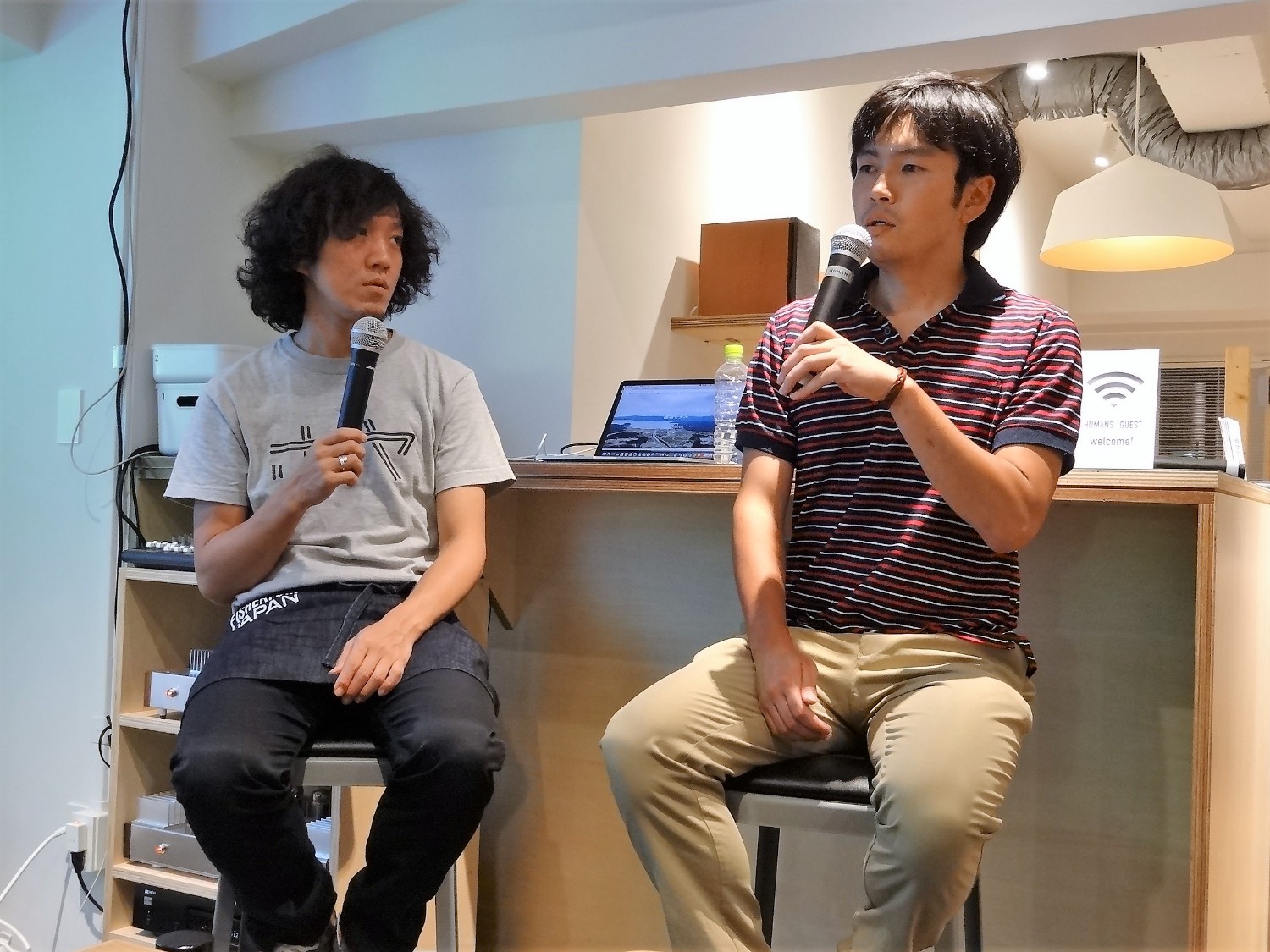 グレーのTシャツを着た長谷川さんと、赤と黒のボーダー柄のポロシャツを着ている正司さんがカウンター前のスツールに座ってマイクを持ちながらトークしている様子の写真