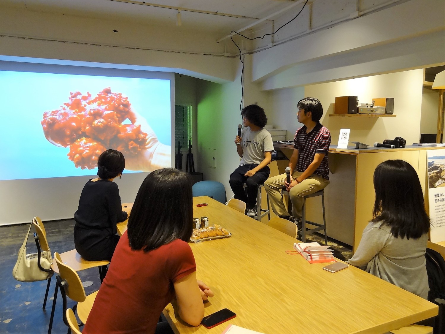 カウンター横の白い壁に赤いホヤの写真が大きく映しだされ、カウンター席に座った長谷川さんがマイクを持って話している様子を、隣の席に座った正司さんと、テーブル席に座った3人の女性が見ている様子の写真